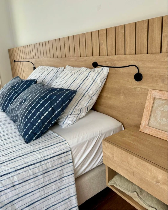 Chambre moderne sous combles avec poutres apparentes blanches, parquet en bois foncé, lit bas contemporain avec coussins gris, fauteuil en rotin blanc, console blanche avec miroir ovale à cadre noir, lampe de table dorée, velux apportant lumière naturelle